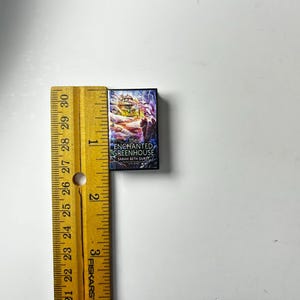 May include: A miniature book titled "The Enchanted Greenhouse" by Sarah Beth Durst, with a colorful cover illustration. The book is next to a yellow ruler, showing its small size. The book's dimensions are approximately 1 inch by 1.5 inches.
