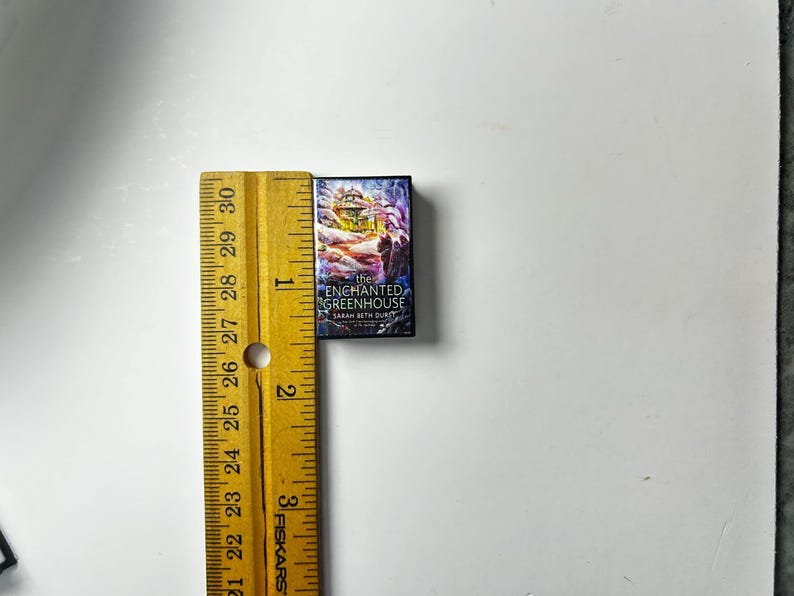 May include: A miniature book titled "The Enchanted Greenhouse" by Sarah Beth Durst, with a colorful cover illustration. The book is next to a yellow ruler, showing its small size. The book's dimensions are approximately 1 inch by 1.5 inches.