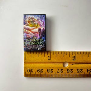 May include: A miniature book titled "The Enchanted Greenhouse" by Sarah Beth Durst. The book cover features a colorful illustration of a building with a purple and blue background. A yellow ruler is next to the book.
