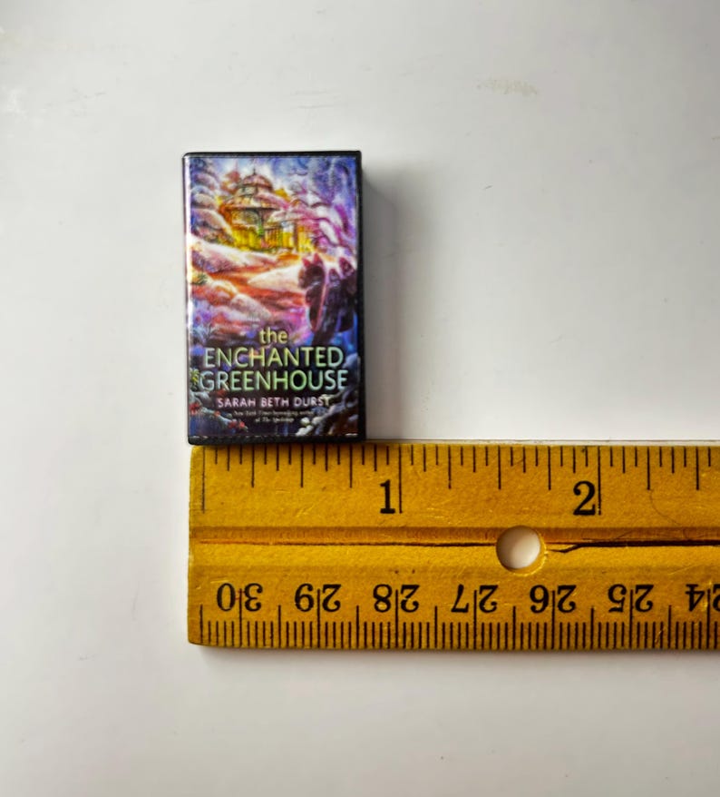 May include: A miniature book titled "The Enchanted Greenhouse" by Sarah Beth Durst. The book cover features a colorful illustration of a building with a purple and blue background. A yellow ruler is next to the book.
