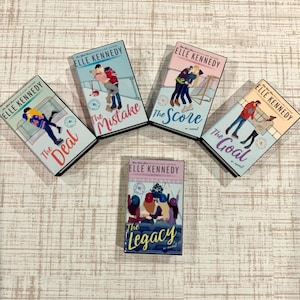 May include: Five books by Elle Kennedy are displayed. The covers feature illustrations of people, hockey players, and the titles "The Deal", "The Mistake", "The Score", "The Goal", and "The Legacy". The books are arranged on a light-colored surface.