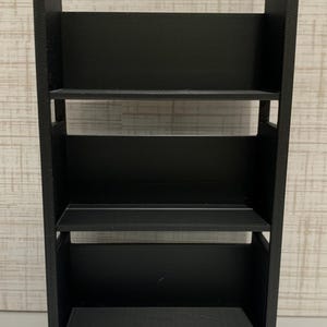 May include: A black three-tiered rolling cart featuring a rectangular frame with two vertical side panels, three horizontal shelves, and two arched handles at the top. The base of the cart is supported by four small wheels attached to the bottom corners.