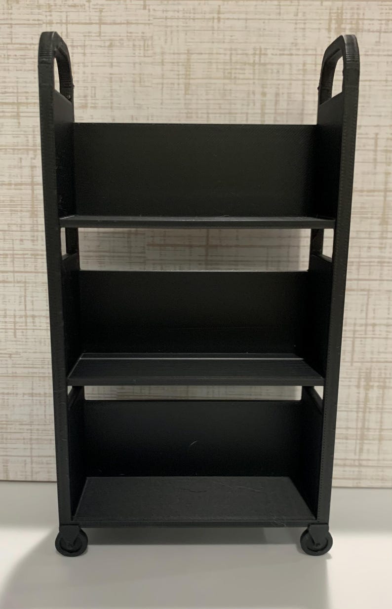 May include: A black three-tiered rolling cart featuring a rectangular frame with two vertical side panels, three horizontal shelves, and two arched handles at the top. The base of the cart is supported by four small wheels attached to the bottom corners.