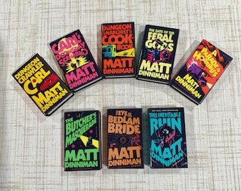 May include: A collection of seven paperback books by Matt Dinniman, including Dungeon Crawler Carl, Carl's Doomsday Scenario, The Dungeon Anarchist's Cook Book, The Gate of the Feral Gods, A Parade of Horribles, The Butcher's Masquerade, and The Eye of the Bedlam Bride.