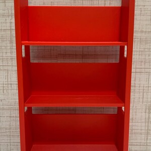 May include: A bright red, three-tiered rolling utility cart featuring a rectangular frame with integrated handles at the top and four small wheels at the base. The cart has three open shelves for storage and a smooth, matte finish throughout.
