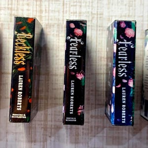 May include: Five hardcover books by Lauren Roberts are displayed. The spines feature titles like "Powerless," "Reckless," and "Fearless," with floral and abstract designs. The books are in various colors, including black, blue, and white.