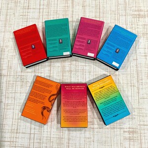 May include: Seven rectangular books arranged in a semi-circle, each featuring a solid color cover including red, green, pink, blue, orange, and a yellow-to-blue gradient. The books display printed text on their back covers.