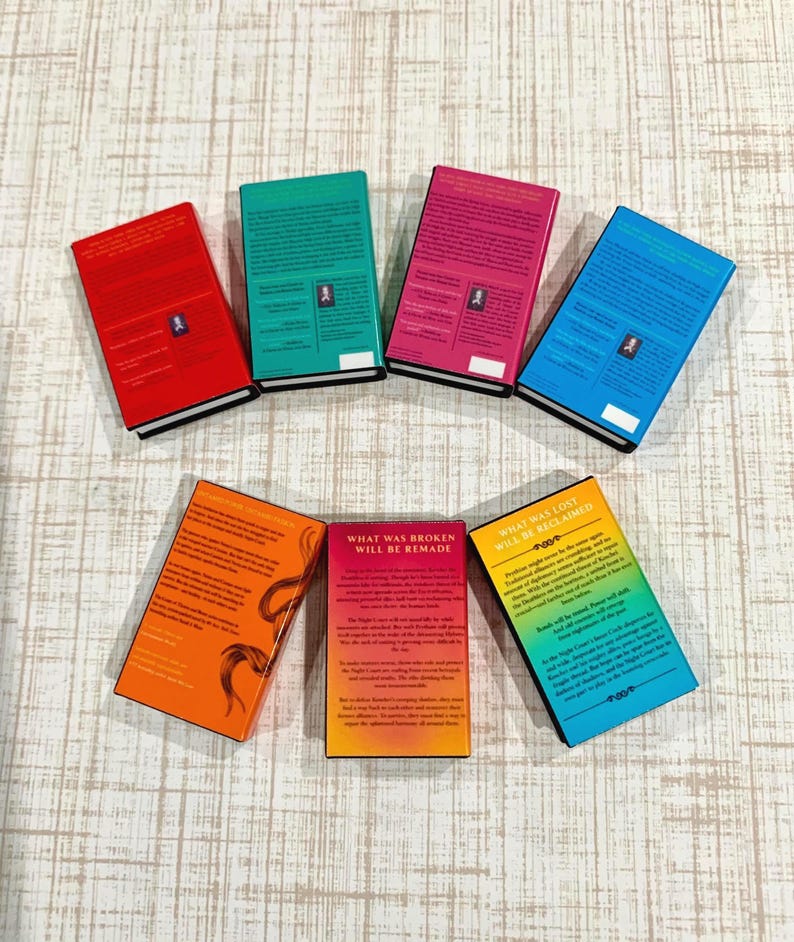 May include: Seven rectangular books arranged in a semi-circle, each featuring a solid color cover including red, green, pink, blue, orange, and a yellow-to-blue gradient. The books display printed text on their back covers.
