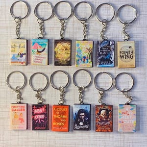 May include: A collection of miniature book keychains. Each keychain features a book cover design with titles like "Fourth Wing" and "A Court of Thorns and Roses." The keychains have silver-colored rings and chains, and the book covers display various colors and illustrations.