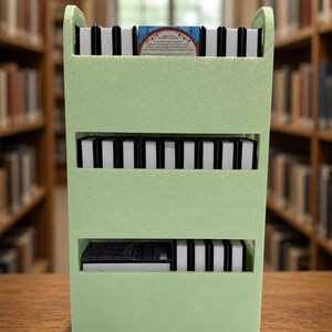 May include: A light green three-tiered storage cart with vertical dividers, holding multiple small rectangular items with black and white spines. The top shelf features one item with visible text and a red circular graphic on its cover.