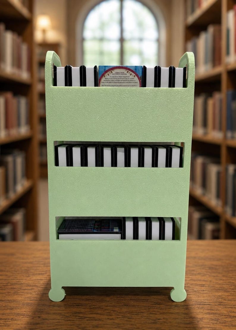 May include: A light green three-tiered storage cart with vertical dividers, holding multiple small rectangular items with black and white spines. The top shelf features one item with visible text and a red circular graphic on its cover.