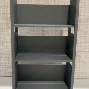 May include: A gray three-tiered rolling cart featuring a tall, rectangular frame with open sides, three flat shelves for storage, and four small wheels at the base. The structure is constructed with vertical side panels and integrated handles at the top.
