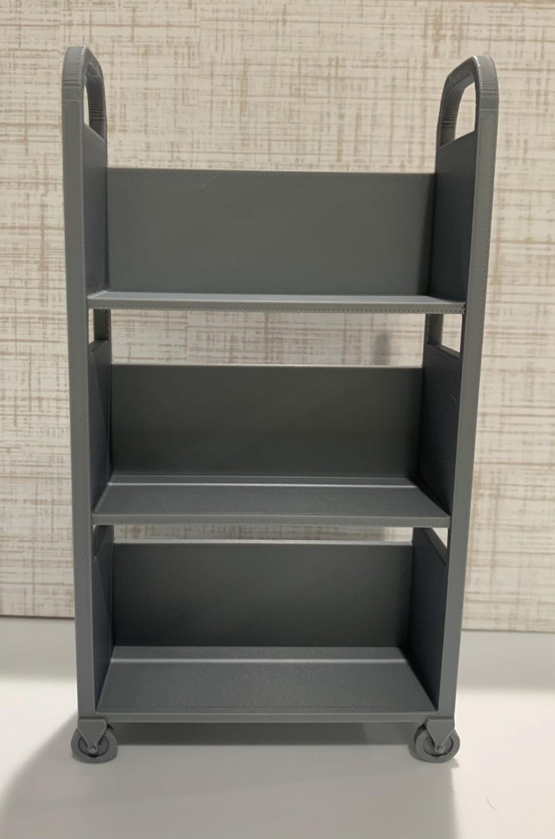 May include: A gray three-tiered rolling cart featuring a tall, rectangular frame with open sides, three flat shelves for storage, and four small wheels at the base. The structure is constructed with vertical side panels and integrated handles at the top.