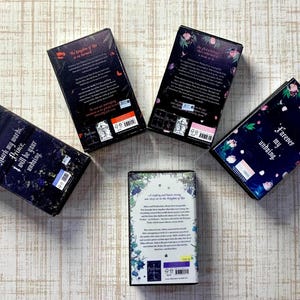 May include: Five hardcover books with dark covers are arranged on a light wood-grain surface. The books have titles and text on the spines and covers. Floral and botanical designs decorate the covers. The books are in a fantasy genre.