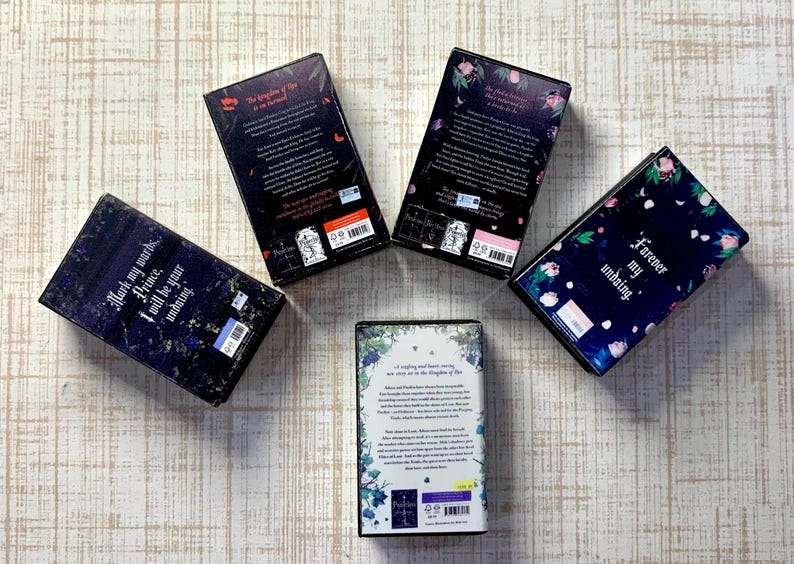 May include: Five hardcover books with dark covers are arranged on a light wood-grain surface. The books have titles and text on the spines and covers. Floral and botanical designs decorate the covers. The books are in a fantasy genre.