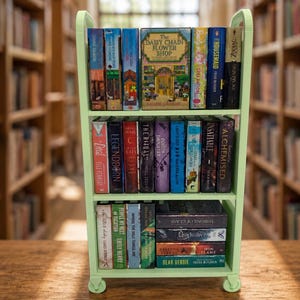 May include: A light green miniature three-tiered rolling cart filled with various small books arranged vertically and horizontally on the shelves. The text *MINI BOOKS NOT INCLUDED* is displayed in red above the cart, which features four small wheels.