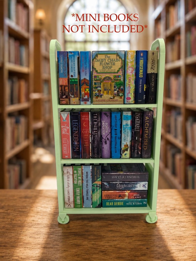 May include: A light green miniature three-tiered rolling cart filled with various small books arranged vertically and horizontally on the shelves. The text *MINI BOOKS NOT INCLUDED* is displayed in red above the cart, which features four small wheels.