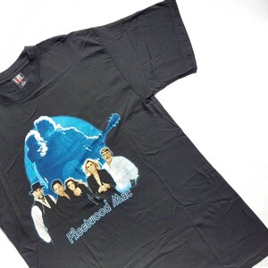 May include: Black t-shirt with a blue silhouette of a person playing guitar and a group photo of Fleetwood Mac band members. The text "Fleetwood Mac" is printed in white below the image.