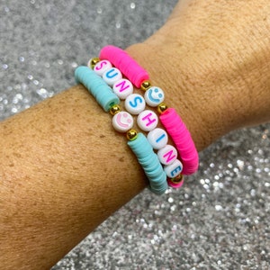 May include: Three colorful beaded bracelets spelling out "SUNSHINE" with smiley face beads. The bracelets are on a person's wrist.