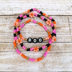 May include: Three layered beaded bracelets in pink, orange, and black with gold accents. The bracelets feature small flower charms and spell out "BOO" in black letters.