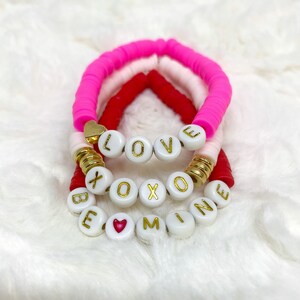 May include: Three colorful silicone bracelets with white beads spelling out "LOVE", "XOXO", and "BE MINE". The beads have gold lettering and a small red heart charm is included.