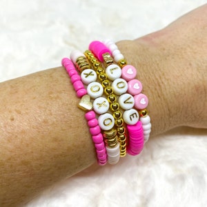 May include: A set of pink and white beaded bracelets with gold beads and letter charms spelling out "LOVE" and "XO".