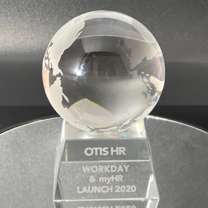 May include: A clear glass globe award with etched continents, mounted on a clear rectangular base. The base features the text "OTIS HR WORKDAY & myHR LAUNCH 2020". The award is displayed on a reflective surface.
