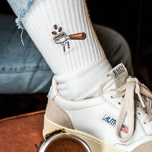 Könnte beinhalten: Weiße Socken mit einem gestickten Kaffeemaschinen-Design. Die Socken werden mit weißen Sneakers mit einem beigefarbenen Wildleder-Panel und dem Wort "AUTH" an der Seite getragen.