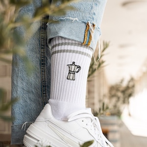 Könnte beinhalten: Weiße Socken mit einem grauen Streifen und einem kleinen gestickten Bild einer Kaffeekanne. Die Socken werden mit weißen Turnschuhen getragen.