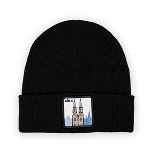 Könnte beinhalten: Schwarze Beanie-Mütze mit einem rechteckigen Aufnäher mit dem Kölner Dom und dem Wort "KÖLN" in Weiß. Der Aufnäher hat einen weißen Rand und einen blau-grauen Hintergrund. Die Mütze ist aus weichem Strickmaterial.