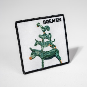 Könnte beinhalten: Ein weißer gestickter Aufnäher mit schwarzen Nähten und dem Wort "BREMEN" in schwarzem Text. Der Aufnäher zeigt ein grünes gesticktes Bild der Bremer Stadtmusikanten, ein Esel, ein Hund, eine Katze und ein Hahn, übereinander gestapelt.
