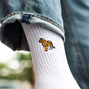 Könnte beinhalten: Weiße gerippte Socken mit einem gestickten Tiger-Design in Orange und Schwarz. Die Socken sind unter einer blauen Jeans zu sehen.