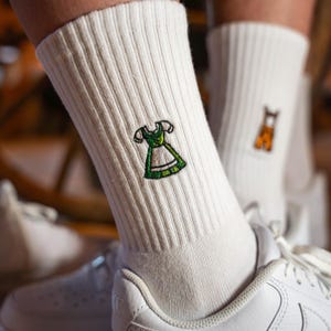 Könnte beinhalten: Weiße Socken mit gestickten Bildern eines traditionellen deutschen Dirndls und einer Lederhose. Die Socken werden an den Füßen einer Person getragen.