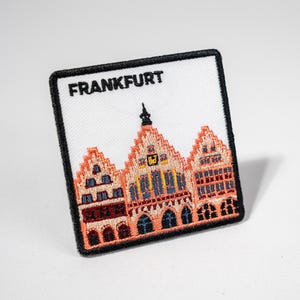 Könnte beinhalten: Ein weißer gestickter Aufnäher mit schwarzen Nähten, der ein rotes Backsteingebäude mit einem Uhrturm und dem Text "FRANKFURT" zeigt.