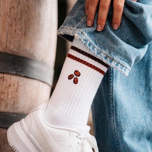 Könnte beinhalten: Weiße Socken mit braunen und weinroten Streifen und gestickten Kaffeebohnen. Die Socken sind gerippt und werden mit weißen Turnschuhen und einer blauen Jeans getragen. Ein lustiges Accessoire.