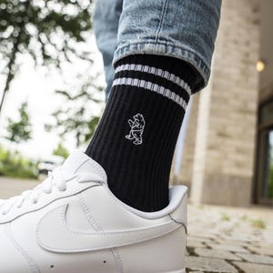 Könnte beinhalten: Schwarze gerippte Socken mit weißen Streifen und einer weißen Bär-Grafik. Die Socken werden mit weißen Turnschuhen und einer blauen Jeans getragen. Die Socken haben zwei weiße Streifen oben.