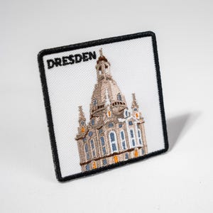 Könnte beinhalten: Weißer gestickter Aufnäher mit schwarzem Rand, der ein detailliertes Bild eines Kirchengebäudes mit einer Kuppel und dem Wort "DRESDEN" in schwarzem Text zeigt.