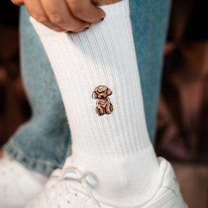 Könnte beinhalten: Weiße gerippte Socken mit einer gestickten Abbildung eines braunen Pudels. Die Socken sind hochgezogen und zeigen das Hunde-Design. Sie werden mit weißen Turnschuhen und einer blauen Jeans getragen.