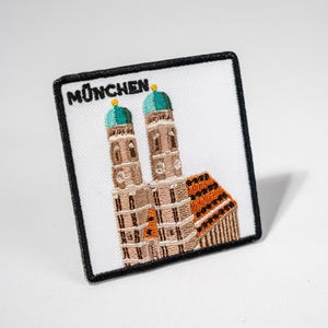 Könnte beinhalten: Ein gestickter Aufnäher mit weißem Hintergrund und schwarzem Rand. Der Aufnäher zeigt ein Gebäude mit zwei Türmen und einem braunen Dach. Der Text "MÜNCHEN" ist oben auf dem Aufnäher in Schwarz gestickt.