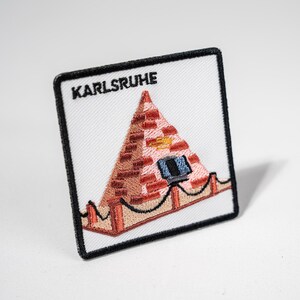 Könnte beinhalten: Gestickter Aufnäher mit einem Pyramiden-Design und dem Wort "KARLSRUHE". Die Pyramide ist aus roten Ziegeln mit einem blauen Fenster und goldenen Details. Der Aufnäher hat einen weißen Hintergrund und einen schwarzen Rand.