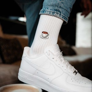 Könnte beinhalten: Weiße gerippte Socken mit einem gestickten Kaffeetassen-Design. Die Socken werden mit weißen Turnschuhen und einer blauen Jeans getragen. Die Kaffeetasse ist braun mit einer weißen Untertasse. Die Turnschuhe haben weiße Schnürsenkel und eine weiße Sohle.
