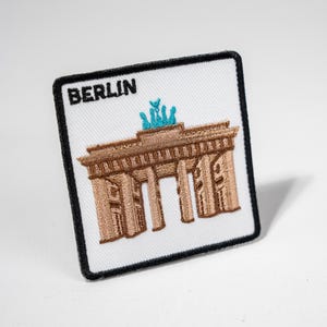 Könnte beinhalten: Ein weißer gestickter Patch mit schwarzem Rand. Der Patch zeigt ein braunes gesticktes Bild des Brandenburger Tors in Berlin, Deutschland. Der Text "BERLIN" ist oben auf dem Patch in Schwarz gestickt.