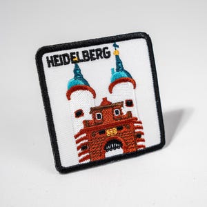 Könnte beinhalten: Weißer gestickter Aufnäher mit dem Wort "HEIDELBERG" in schwarzen Buchstaben und einem Bild eines roten Backsteingebäudes mit zwei weißen Türmen, die mit blauen und weißen Türmen gekrönt sind.