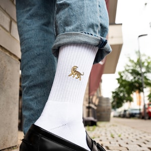 Könnte beinhalten: Weiße gerippte Crew-Socken mit einem kleinen, gestickten Leopardenmuster in Beige- und Brauntönen. Die Socken werden mit blauen Jeans und schwarzen Lederloafers getragen.