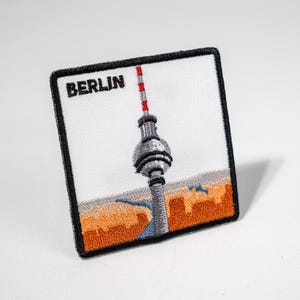 Könnte beinhalten: Gestickter Aufnäher mit dem Berliner Fernsehturm und dem Wort "BERLIN" oben. Das Design zeigt den Turm in Grau, Rot und Weiß, vor einem weißen Hintergrund mit orangefarbenen und grauen Akzenten. Der Aufnäher hat einen schwarzen Rand.