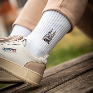 Könnte beinhalten: Weiße gerippte Socken mit einem farbenfrohen, gestickten Kathedralen-Design. Die Socken werden mit cremefarbenen Turnschuhen mit beigem Wildlederakzent und einem kleinen amerikanischen Flaggenaufnäher getragen.