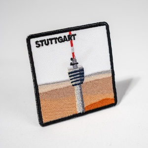 Könnte beinhalten: Ein weißer gestickter Aufnäher mit schwarzen Nähten, der eine stilisierte Illustration des Stuttgarter Fernsehturms mit dem Text "STUTTGART" darüber zeigt.