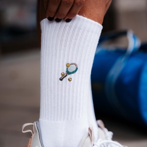 Könnte beinhalten: Weiße Sportsocken mit gerippter Struktur, mit einer gestickten Tennis-Schlägern und zwei Tennisbällen in Grün-, Braun- und Gelbtönen. Die Socken werden mit weißen und rosa Turnschuhen getragen.