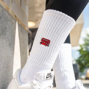Könnte beinhalten: Weiße gerippte Socken mit einem roten Doppeldeckerbus, der auf der Seite gestickt ist. Die Socken werden mit weißen Turnschuhen getragen. Das Bild ist im Freien aufgenommen.