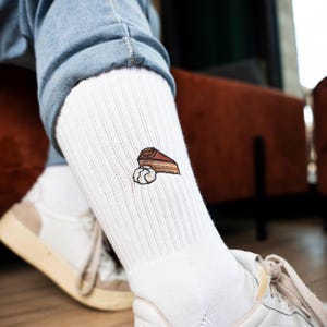 Könnte beinhalten: Weiße gerippte Socken mit einer gestickten Darstellung eines Kuchenstücks und Schlagsahne. Die Socken werden mit blauen Jeans und weißen Turnschuhen getragen.